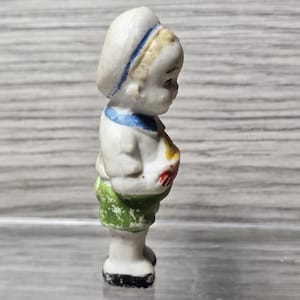 Pu&ograve; includere: Piccola statuina vintage in porcellana di un bambino in tenuta da marinaio. La statuina ha un cappello bianco con una striscia blu, un colletto blu e pantaloncini verdi. Il bambino tiene in mano un oggetto rosso e giallo.