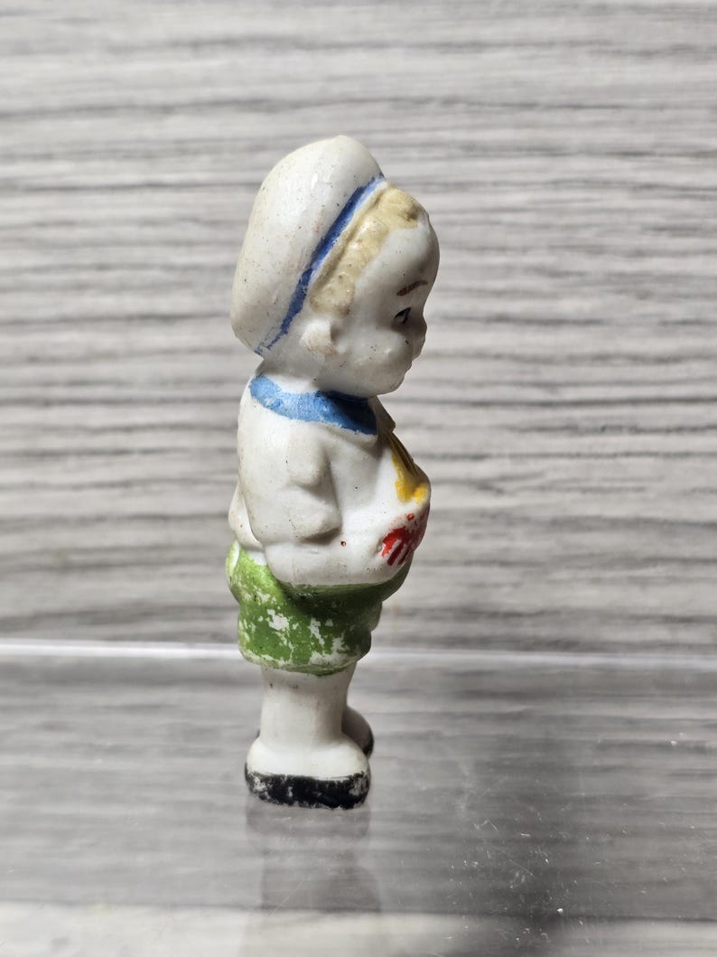 Pu&ograve; includere: Piccola statuina vintage in porcellana di un bambino in tenuta da marinaio. La statuina ha un cappello bianco con una striscia blu, un colletto blu e pantaloncini verdi. Il bambino tiene in mano un oggetto rosso e giallo.