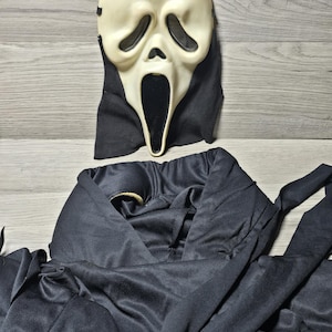 May include: A white plastic ghost mask with large black eye openings and an elongated black mouth opening, attached to a black fabric hood. Below the mask is a separate, large black fabric robe with long sleeves and a wide, draped collar.