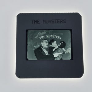 May include: A black and white slide featuring a scene from "The Munsters" television show. The slide shows three characters, with the show's title at the top and on the image. The image is set against a white background.