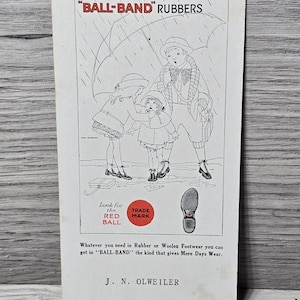 May include: Vintage advertisement for "BALL-BAND" rubbers. The black and white illustration depicts three children under an umbrella in the rain. The text includes "Look for the RED BALL" and "TRADE MARK". The advertisement promotes rubber and woolen footwear.