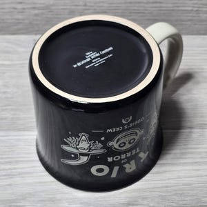 Puede incluir: Taza de cer&aacute;mica negra con asa y borde blancos, con personajes de "Pesadilla antes de Navidad". El dise&ntilde;o incluye las palabras "RIO OF TERROR" y "Oogie's Crew."