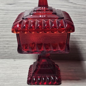 May include: A ruby red glass candy dish with a lid. The dish has a square base and a pedestal. The lid has a small handle on top. The glass has a textured, faceted design.