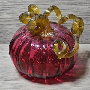 Può includere: Una zucca decorativa in vetro con un corpo rosso vibrante e un gambo dorato e a spirale. La superficie della zucca è strutturata con creste verticali e il gambo è finemente progettato. Questo articolo a tema autunnale è perfetto per l'arredamento della casa.