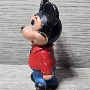 May include: A vintage Mickey Mouse figurine. The figure is wearing a red jacket, blue trousers, and black shoes. The mouse has black ears and a tan face. The figurine is standing and facing left.
