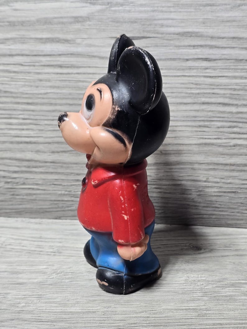 May include: A vintage Mickey Mouse figurine. The figure is wearing a red jacket, blue trousers, and black shoes. The mouse has black ears and a tan face. The figurine is standing and facing left.