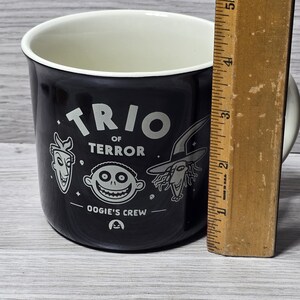 Puede incluir: Taza de cer&aacute;mica negra con interior blanco. La taza presenta el texto "TRIO of TERROR" e ilustraciones de personajes de una pel&iacute;cula. La taza mide aproximadamente 10 cm de alto.