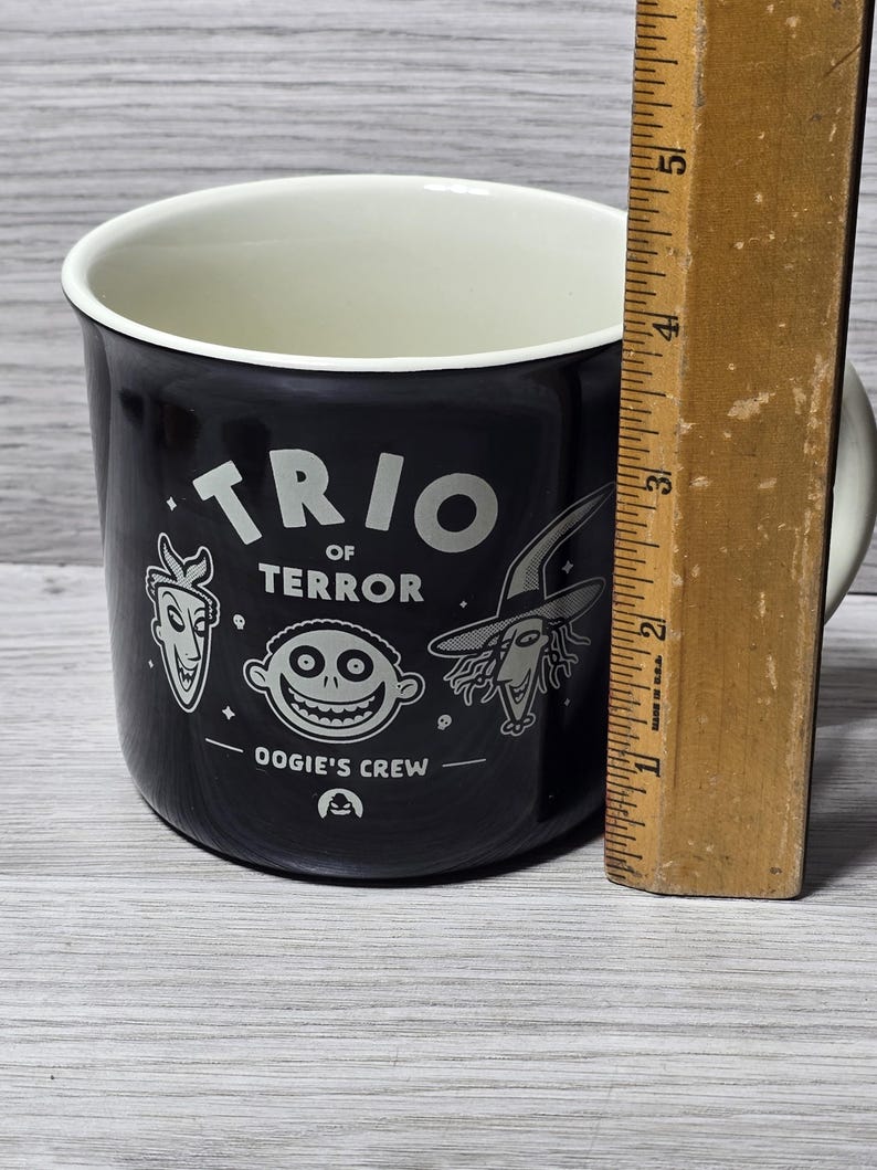 Puede incluir: Taza de cer&aacute;mica negra con interior blanco. La taza presenta el texto "TRIO of TERROR" e ilustraciones de personajes de una pel&iacute;cula. La taza mide aproximadamente 10 cm de alto.