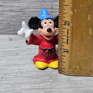 May include: A small, colorful figurine of Mickey Mouse dressed as a sorcerer, with a blue hat and red robe. The figure is approximately 2 inches tall. The figure is standing next to a wooden ruler.