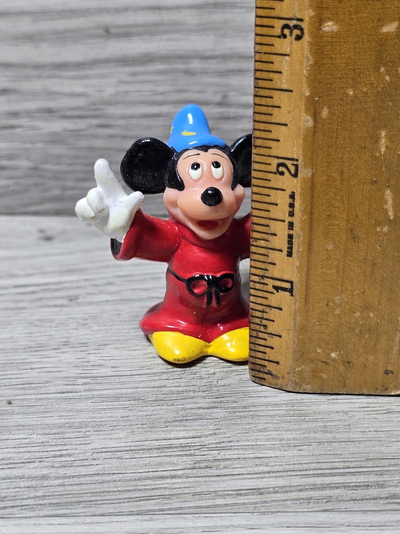 May include: A small, colorful figurine of Mickey Mouse dressed as a sorcerer, with a blue hat and red robe. The figure is approximately 2 inches tall. The figure is standing next to a wooden ruler.