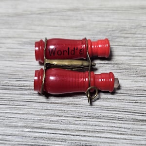 Könnte beinhalten: Zwei rote Flaschenverschlüsse im Vintage-Stil mit dem Aufdruck "World's". Die Verschlüsse werden von einem Metallverschluss und Drahtringen zusammengehalten. Der Hintergrund ist ein hellgraues und weißes Holzmaserungsmuster.