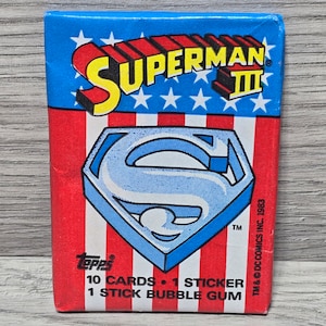May include: A red, white, and blue card pack for Superman III trading cards. The pack features a blue and white Superman logo with the text "SUPERMAN III" in red and white letters. The pack includes 10 cards, 1 sticker, and 1 stick of bubble gum.