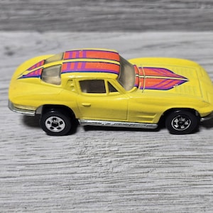 Op de afbeelding: Een gele speelgoedauto met een kleurrijk ontwerp op het dak en de motorkap. De auto heeft zwarte wielen en een zilveren afwerking. Het ontwerp omvat oranje, paarse en rode strepen en vormen. Deze vintage speelgoedauto is een verzamelobject.