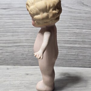 May include: A vintage bisque doll figurine, nude with a light skin tone. The doll has a detailed, curly blonde hairstyle and is standing upright. The doll's arms are slightly outstretched, and the figure is on a gray wooden surface.