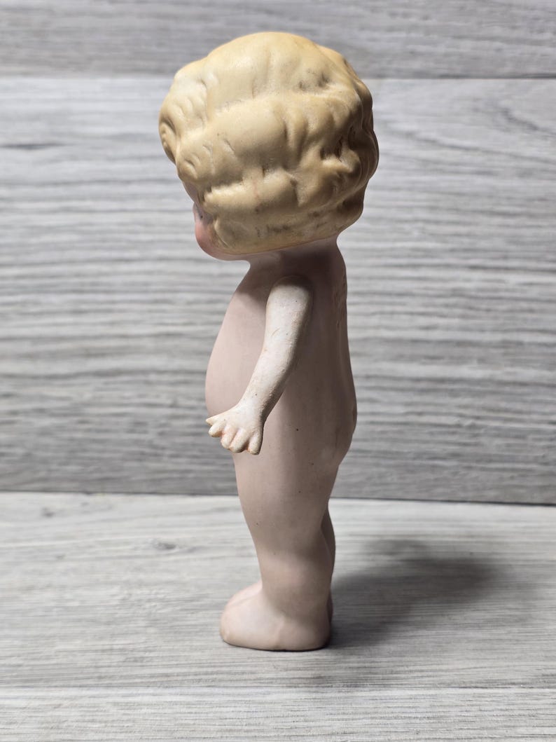 May include: A vintage bisque doll figurine, nude with a light skin tone. The doll has a detailed, curly blonde hairstyle and is standing upright. The doll's arms are slightly outstretched, and the figure is on a gray wooden surface.