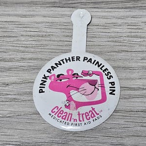May include: A white round pin with a pink cartoon panther wearing a red bow tie. The pin says "Pink Panther Painless Pin" and "clean 'n treat Medicated First Aid Pads" in black text.