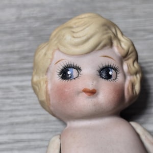 May include: Close-up of a vintage porcelain doll head. The doll has blonde hair, blue eyes with long eyelashes, and rosy cheeks. The doll's lips are a soft peach color. The doll's skin is a pale, porcelain color.