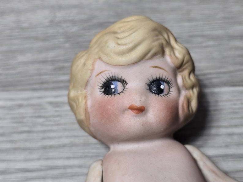 May include: Close-up of a vintage porcelain doll head. The doll has blonde hair, blue eyes with long eyelashes, and rosy cheeks. The doll's lips are a soft peach color. The doll's skin is a pale, porcelain color.