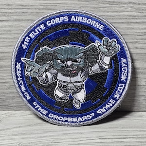 Könnte beinhalten: Ein kreisförmiger Aufnäher mit blauem Rand und dem Text "41st ELITE CORPS AIRBORNE" und "THE DROPBEARS". Der Aufnäher zeigt einen Cartoon-Drop-Bear-Charakter in weißer und grauer Uniform mit schwarzem Hintergrund.