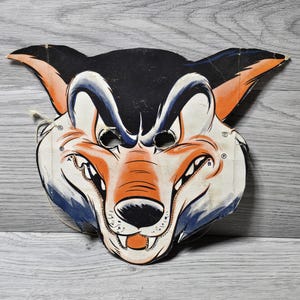 Peut inclure: Masque en papier vintage représentant un visage de loup ou de renard stylisé. Le masque a un dessus noir, un visage orange et blanc avec des accents bleus et des trous pour les yeux. Il a une ficelle pour le porter et est probablement un costume d'Halloween.