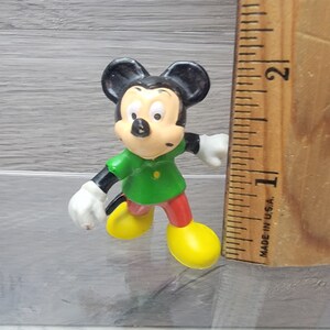 Pode incluir: Uma pequena figura de pl&aacute;stico do Mickey Mouse vestindo uma camisa verde e cal&ccedil;as vermelhas. Ele est&aacute; em p&eacute; em uma superf&iacute;cie de madeira com uma r&eacute;gua ao lado. A r&eacute;gua &eacute; marcada em polegadas e diz "Made in U.S.A."