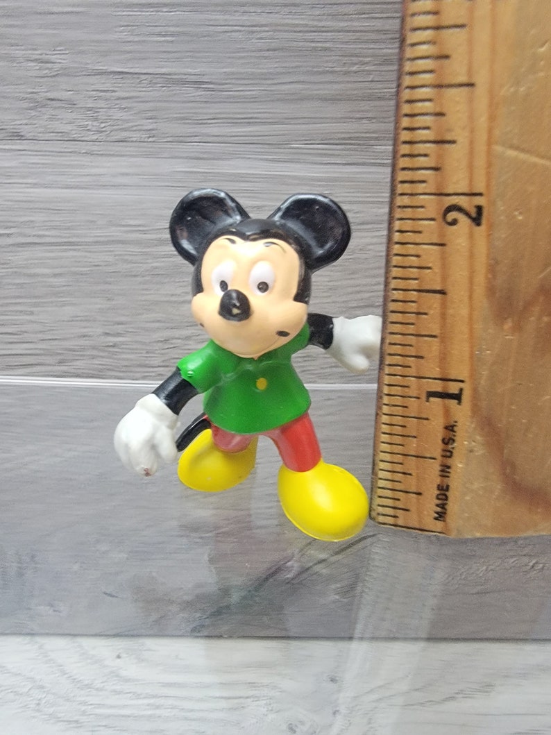 Pode incluir: Uma pequena figura de pl&aacute;stico do Mickey Mouse vestindo uma camisa verde e cal&ccedil;as vermelhas. Ele est&aacute; em p&eacute; em uma superf&iacute;cie de madeira com uma r&eacute;gua ao lado. A r&eacute;gua &eacute; marcada em polegadas e diz "Made in U.S.A."