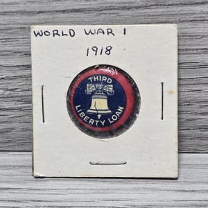May include: A circular pinback button featuring a blue center with a cream-colored Liberty Bell graphic and the text THIRD LIBERTY LOAN. The button has a red border and is encased in a square white cardboard holder labeled WORLD WAR I 1918.