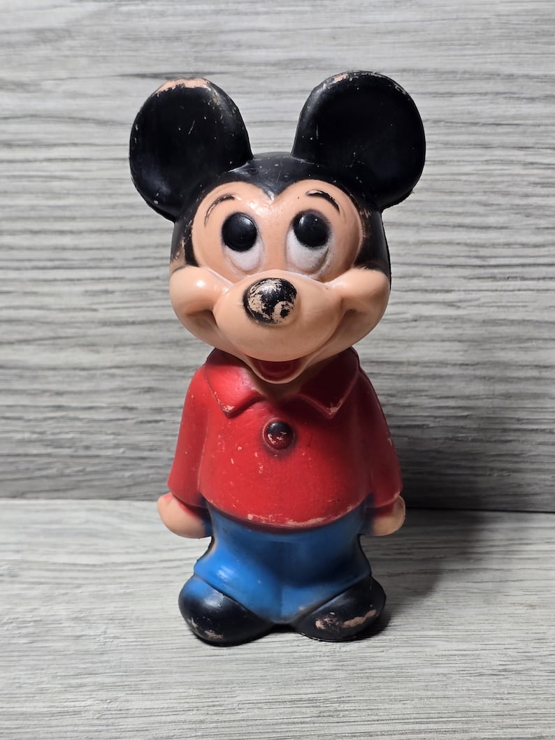 May include: A vintage Mickey Mouse figure, posed in a classic stance. The figure has black ears, a tan face, and wears a red shirt and blue shorts. The toy shows signs of wear, adding to its vintage charm.