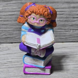 May include: A figurine of a girl with red hair in pigtails wearing glasses and purple overalls, reading a book. She is sitting on a stack of three books with blue and pink covers. The figurine has a whimsical, cartoonish style.