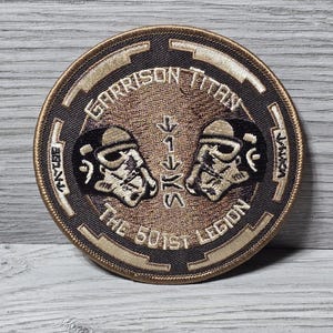 以下が含まれることがあります： 茶色と金色のデザインの円形パッチ。2つのストームトルーパーのヘルメットが向かい合っており、金色の文字で「GARRISON TITAN」と「THE 501ST LEGION」と書かれています。このパッチはおそらく収集品です。