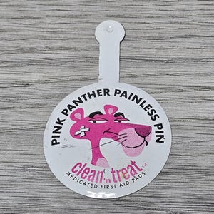 Könnte beinhalten: Ein runder weißer Knopf mit einer pinkfarbenen Comic-Panther mit einem Verband auf der Nase. Der Knopf trägt die Aufschrift "Pink Panther Painless Pin" und "clean'n treat Medicated First Aid Pads".