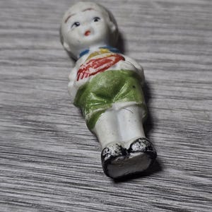 Pu&ograve; includere: Una piccola bambola vintage in porcellana di un ragazzo. La bambola ha un viso bianco, capelli corti e indossa un colletto blu, un gilet rosso e giallo e pantaloncini verdi. Le scarpe della bambola sono bianche e nere.
