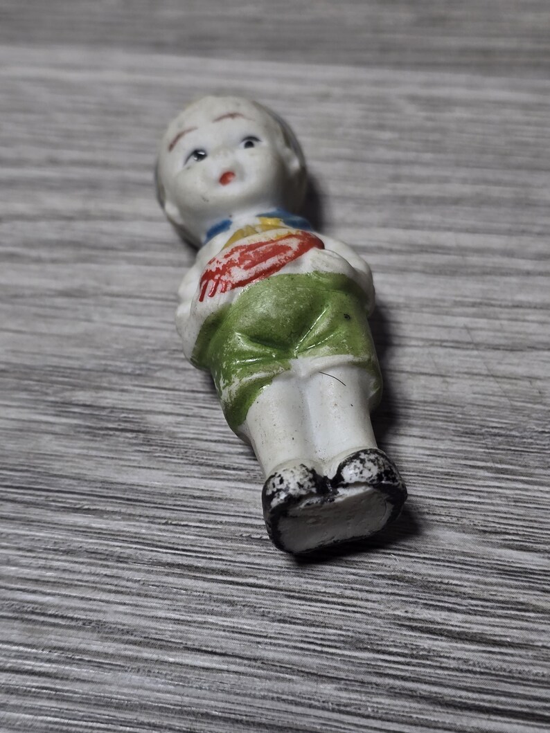 Pu&ograve; includere: Una piccola bambola vintage in porcellana di un ragazzo. La bambola ha un viso bianco, capelli corti e indossa un colletto blu, un gilet rosso e giallo e pantaloncini verdi. Le scarpe della bambola sono bianche e nere.