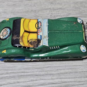 May include: A vintage green tin toy car featuring yellow seats, a black steering wheel, and a circular emblem on the hood. The vehicle has silver-colored bumpers at the front and rear, with visible signs of wear and oxidation on the metal surface.