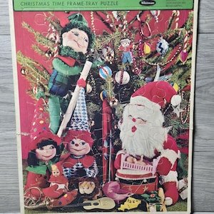 Op de afbeelding: Vintage kerstframe-tray puzzel met de kerstman, elfen en een versierde kerstboom. De puzzel heeft een rode achtergrond en de tekst "Christmas Time Frame-Tray Puzzle" en "Whitman".