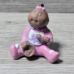 May include: A vintage Cabbage Patch Kids figurine. The doll is seated, wearing a pink outfit with a white bib featuring a purple elephant. The doll has brown skin, a pink bow, and is holding a small bowl.