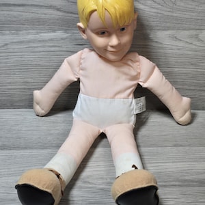 May include: A vintage doll with a painted face, yellow hair, and a soft body. The doll is wearing a pink shirt, white diaper, and has tan and black shoes. The doll's arms and legs are outstretched.