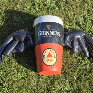 May include: A black inflatable beer stein with wings. The stein is decorated with the Guinness logo and the text "Draught". The wings are black and have a textured surface. The stein is sitting on a green grassy surface.