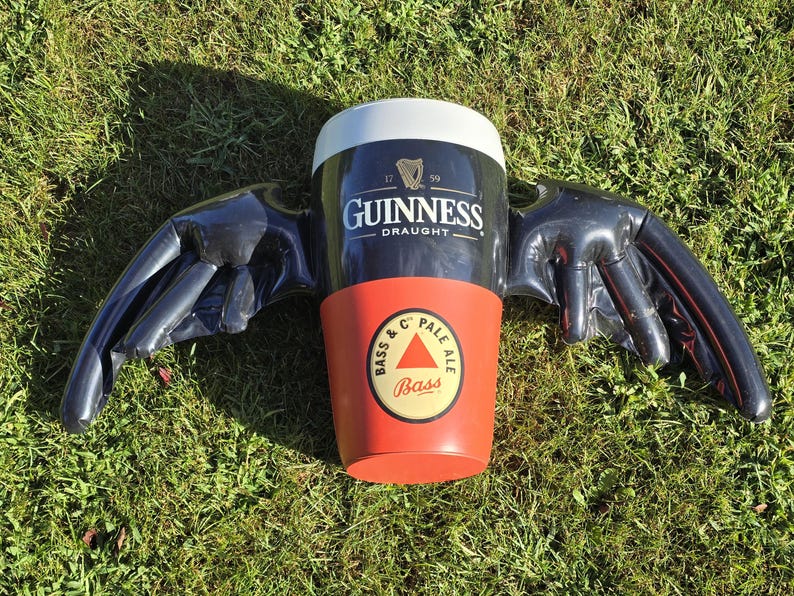 May include: A black inflatable beer stein with wings. The stein is decorated with the Guinness logo and the text "Draught". The wings are black and have a textured surface. The stein is sitting on a green grassy surface.