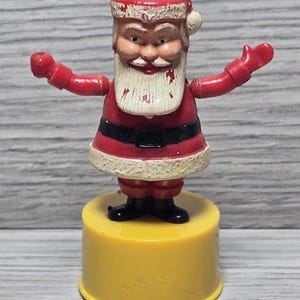 May include: A vintage Santa Claus figurine with a red suit, white beard, and black belt, standing on a round yellow plastic base. The figure has arms extended outward, with visible paint wear on the hat, beard, and suit, showing a weathered texture.