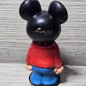 May include: Vintage Mickey Mouse figure. The black-eared figure is dressed in a red top and blue trousers. The toy shows signs of wear, indicating it's an older collectible. The figurine is standing upright.