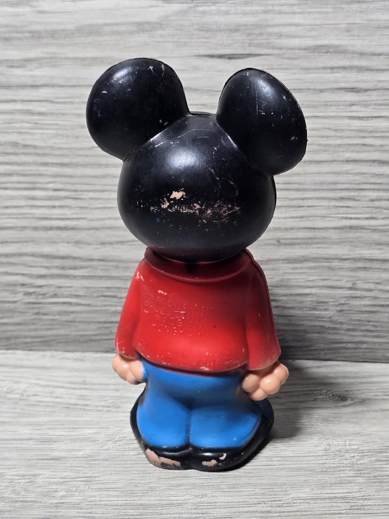 May include: Vintage Mickey Mouse figure. The black-eared figure is dressed in a red top and blue trousers. The toy shows signs of wear, indicating it's an older collectible. The figurine is standing upright.