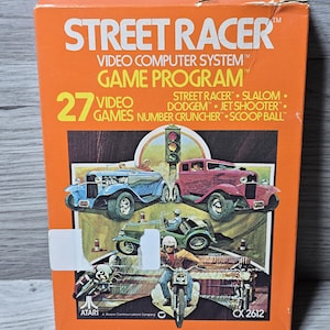 Peut inclure: Boîte orange du jeu vidéo Atari Street Racer. La boîte présente le titre du jeu, une liste de 27 jeux vidéo et des illustrations de voitures et de motos. Le logo Atari et le numéro de modèle CX 2612 sont également visibles.