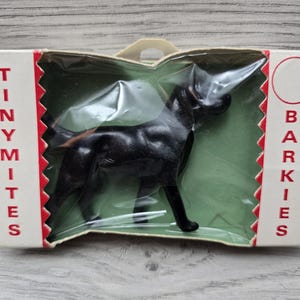 Può includere: Un cane giocattolo di plastica nera in una confezione di plastica trasparente con un retro in cartone rosso e bianco. La confezione dice "Tiny Mites Barkies".