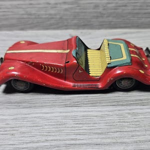 Pode incluir: Um carro de brinquedo de folha de flandres vermelho vintage com um capô longo, uma faixa creme no centro e um banco amarelo com nervuras. O veículo aberto tem uma cobertura traseira azul-petróleo, para-choques pretos e rodas raiadas cinza-escuras.