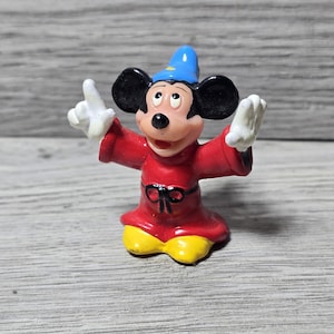 May include: A figurine of Mickey Mouse dressed as a sorcerer, wearing a red robe with a black bow, yellow shoes, and a blue hat. The figure has white gloves and is posed with arms outstretched.