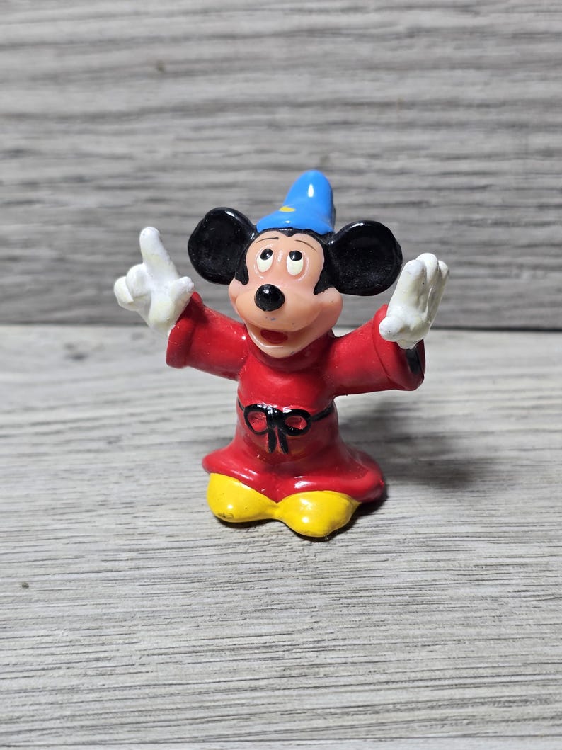May include: A figurine of Mickey Mouse dressed as a sorcerer, wearing a red robe with a black bow, yellow shoes, and a blue hat. The figure has white gloves and is posed with arms outstretched.