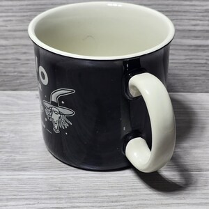 Puede incluir: Taza de cer&aacute;mica negra con interior y asa blancos. La taza presenta un dibujo de bruja de dibujos animados y la palabra "Hello" en blanco. El asa es curvada y de dise&ntilde;o s&oacute;lido.