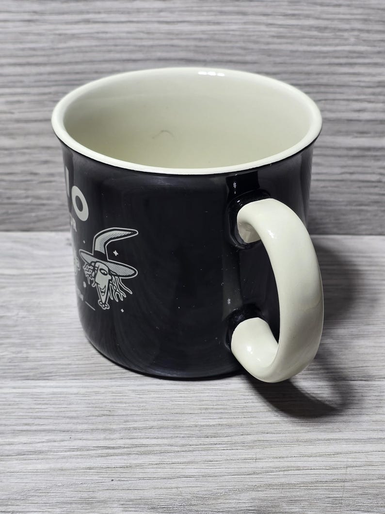 Puede incluir: Taza de cer&aacute;mica negra con interior y asa blancos. La taza presenta un dibujo de bruja de dibujos animados y la palabra "Hello" en blanco. El asa es curvada y de dise&ntilde;o s&oacute;lido.