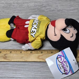 K&ouml;nnte beinhalten: Ein gelbes, rotes und schwarzes Pl&uuml;sch-Mickey-Mouse-Spielzeug. Das Spielzeug tr&auml;gt ein gelbes Shirt mit dem Text "Disney Store" und "Mickey" darauf. Das Spielzeug liegt auf einer Holzoberfl&auml;che.